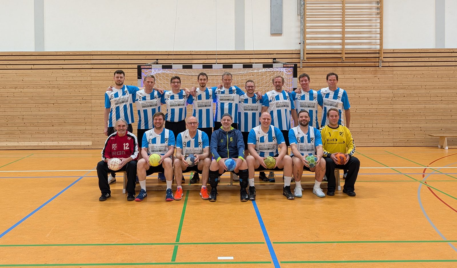 Die Herrenmannschaft von DJK Komet in blau-weiß gestreiften Trikots vor einem Handballtor in der Halle